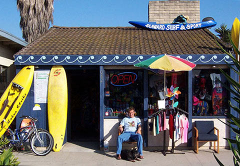 Storefront of a Surf Shop at Ventura, CA Beach with a Kayak sign and surfboards, T-shirts, Sweatshirts, and beach cruiser bicycles out front. 